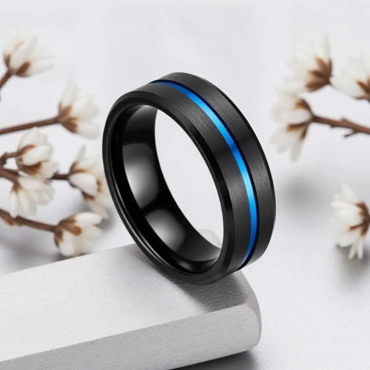 The Visionary black men's wedding ring displayed upright against a white background with white flowers, showcasing its modern brushed black finish.
