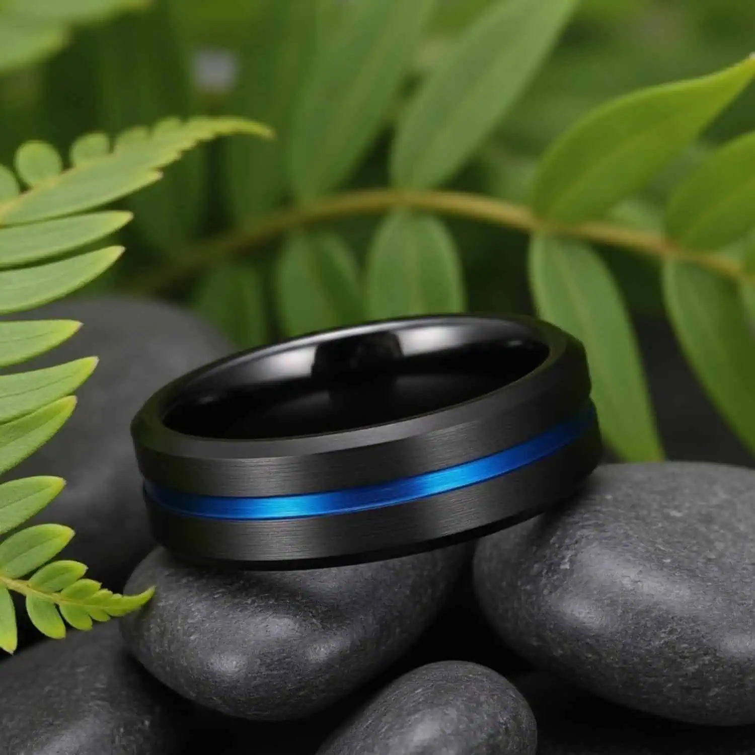 Side view of the Visionary black tungsten ring resting on natural pebbles with lush green leaves softly blurred in the background, showcasing its brushed finish, blue centre groove, and polished bevelled edges.