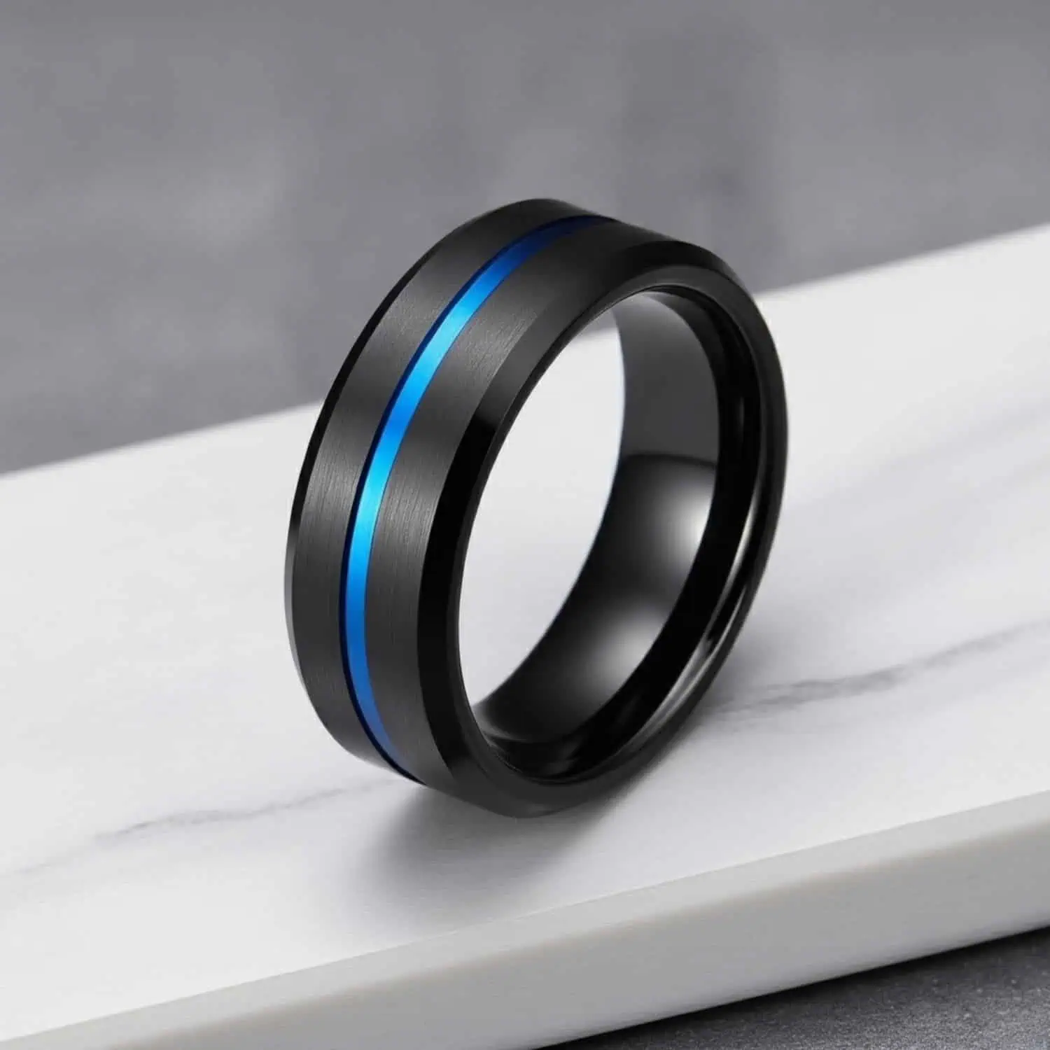 Macro photograph of the Visionary tungsten ring standing on white marble, highlighting the brushed black finish, blue centre groove, and polished bevelled edges.