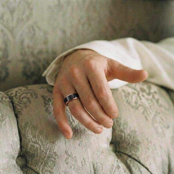 Relaxing on a sofa, you can just see a mans arm and hand. Wearing a white shirt, he is wearing a tungsten ring on this wedding finger. It is a black and rose gold ring.