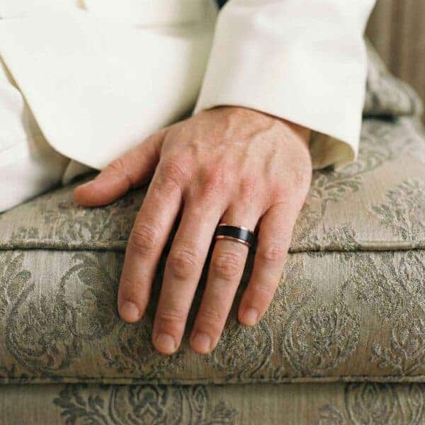 Sitting on a sofa, this is a close up image of a mans hand. You can see that the man is wearing a white suit. The ring is a black and rose gold band made with tungsten.