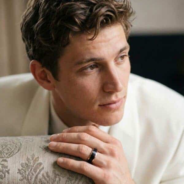 A young man wearing a white suit looks off to one side of the camera. He is wearing a black and rose gold tungsten ring on his wedding finger.