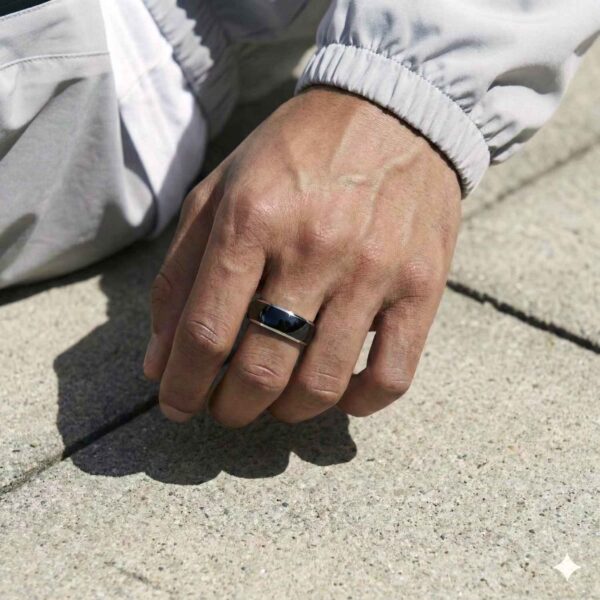 Sitting on the ground, this is a close up shot of a mans hand. On his middle finger, he is wearing a black dome shaped tungsten ring that has polished silver edges.