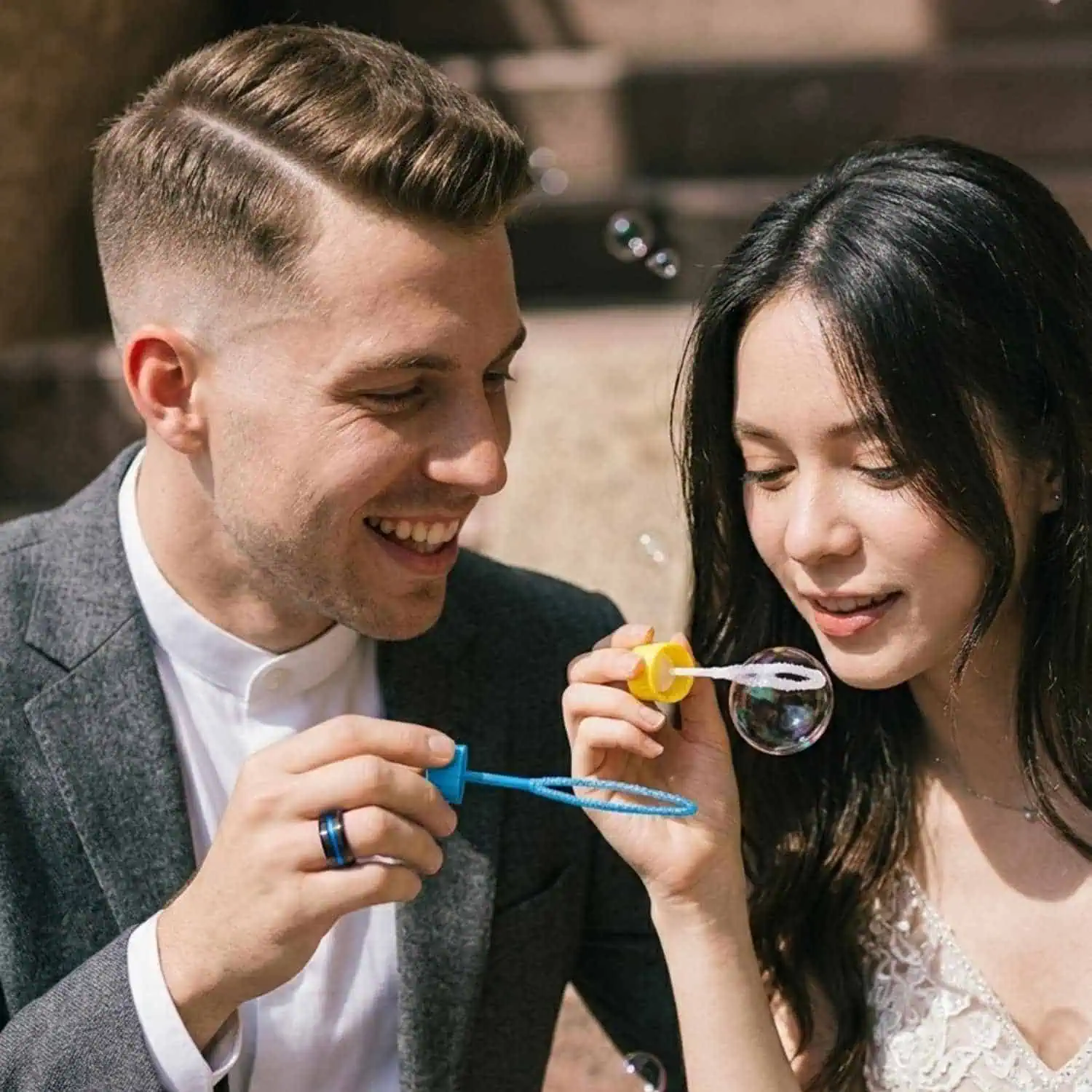 Bride and groom sitting together outside a church blowing bubbles, with the groom wearing the Visionary brushed black tungsten wedding ring featuring a blue centre groove.