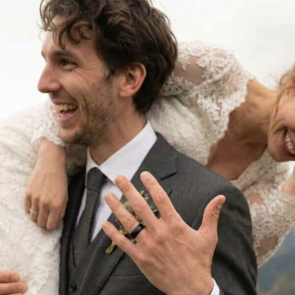 Depicting a happy couple on their wedding day. Dressed in his wedding suit, the man carries his new wife. He holds up his hand showing his black wedding band. It has a domed profile and a lightly brushed finish. The inside of the ring is polished blue.