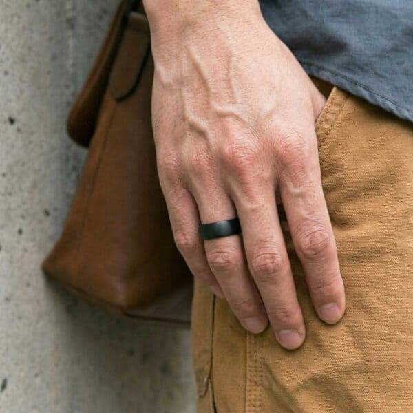 A man rests his hand above his pocket. On his hand, he wears a black ring on his wedding finger. It is a lightly brushed black ring with a domed profile. The inside of the ring is blue.