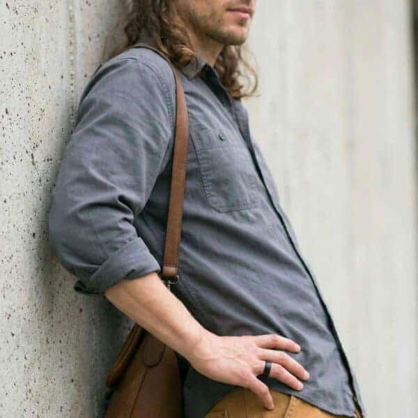 A man rests his back against the wall. With a bag over his shoulder and looking relaxed, he has his hands on his hips. He is wearing a black tungsten ring. The ring is blue on the inside and has a brushed finish.