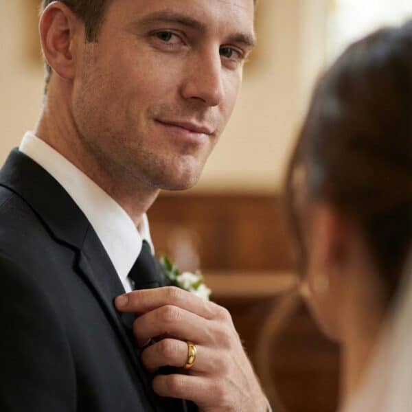 The newly married couple stand together during the wedding ceremony. The man looks into the camera with a smile on his face. He has his hand raised up so that you can see his polished gold tungsten wedding band.