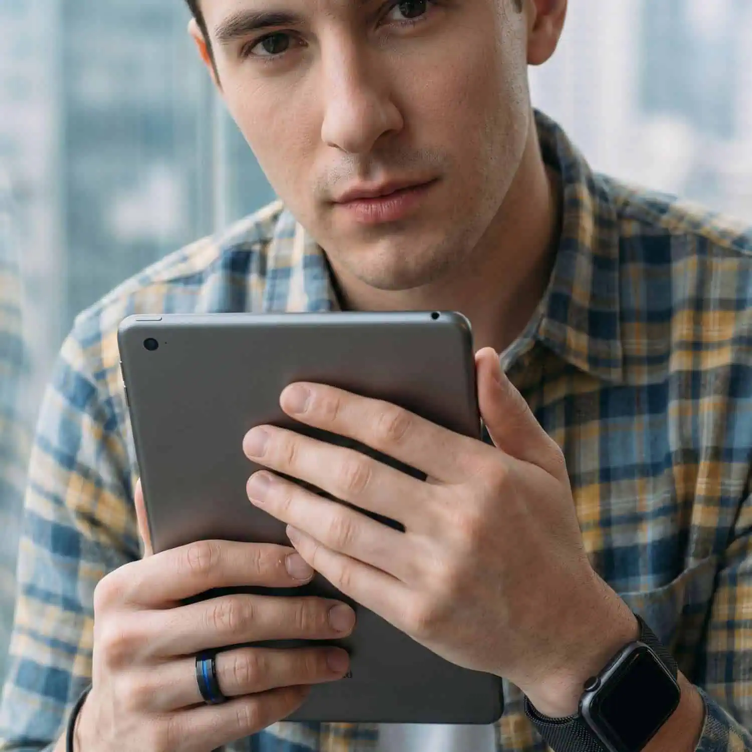 Man wearing a checked shirt holding an iPad while wearing the Visionary men's black tungsten ring with a brushed finish, blue centre groove, and polished black bevelled edges.