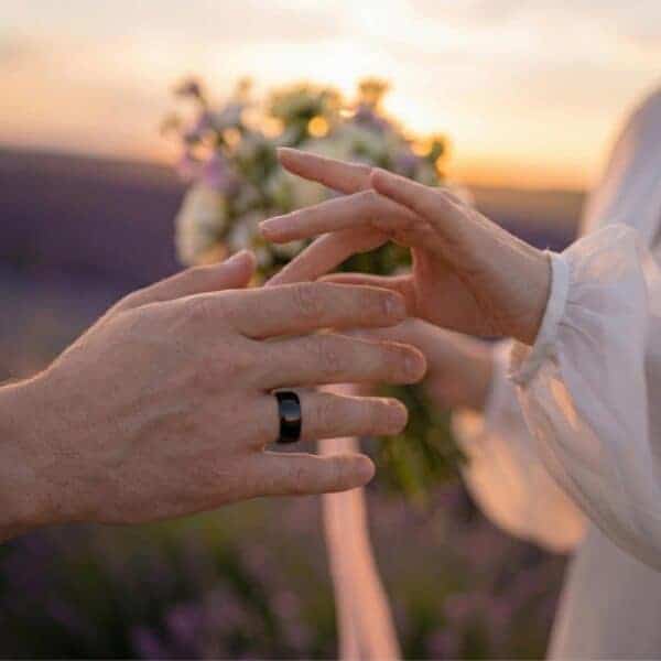 A close up view of the wedding couple's hands. While holding a bouquet of flowers, the bride reaches for her mans hand. As his wedding ring, he wears a polished black tungsten band.
