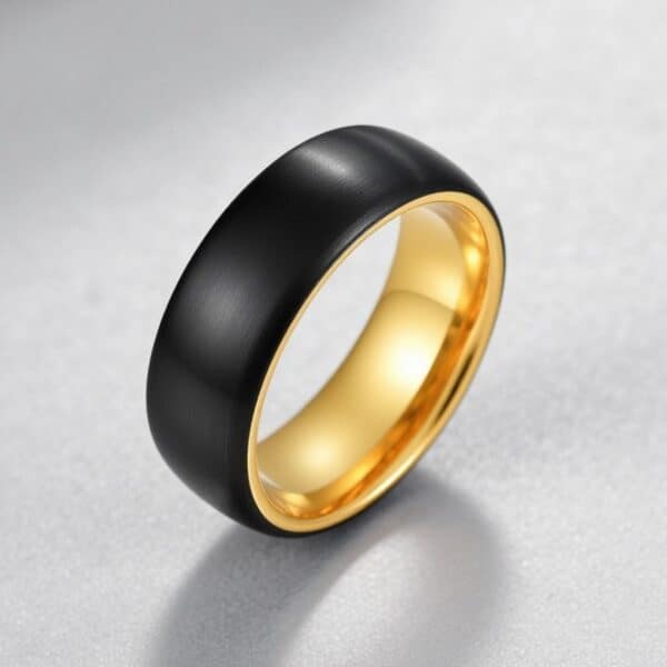 On a light grey surface, a ring stands upright. It is a black tungsten ring with a lustrous gold inside. The ring is slightly domed in shape