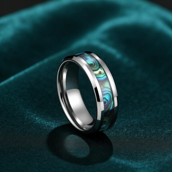 A close up image of a mens ring with abalone shell. Set on a dark green cloth, the ring stands upright. It is a silver tungsten ring featuring turquoise coloured shell.
