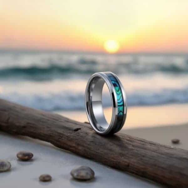 The sun is setting over the rolling waves. Driftwood has been washed up onto the shore. On the driftwood, the ring stands upright. It is a silver tungsten ring with an inlay of shimmering abalone shell.