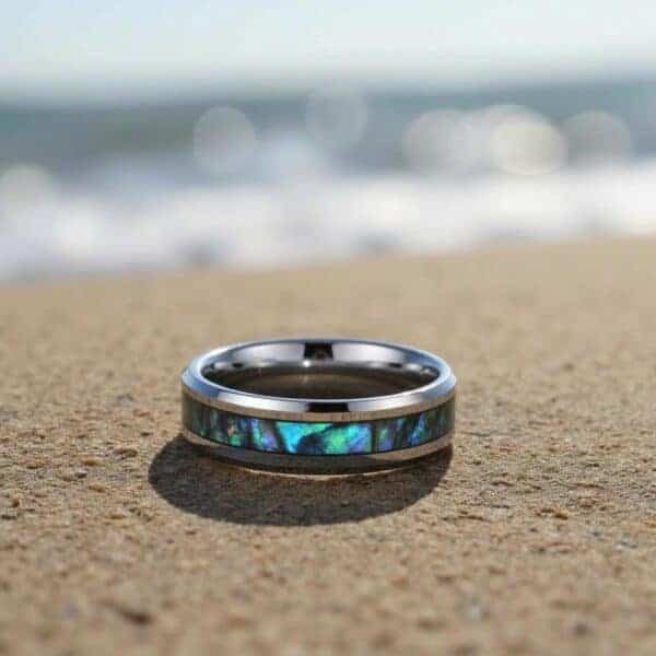 With the sand, sea, and sky above it, a ring lays on a sandy beach. This close up shot of the ring shows a silver tungsten ring with an inlay of abalone shell.
