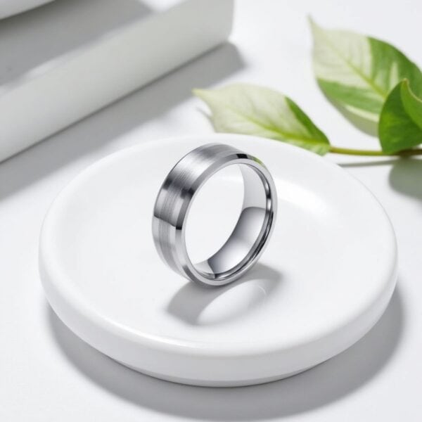 Standing upright on a white stand, and with a white background, sits a silver tungsten ring. With green leaves in the background, this silver tungsten ring features bevelled edges and a contrasting brushed silver section at the middle of the ring.