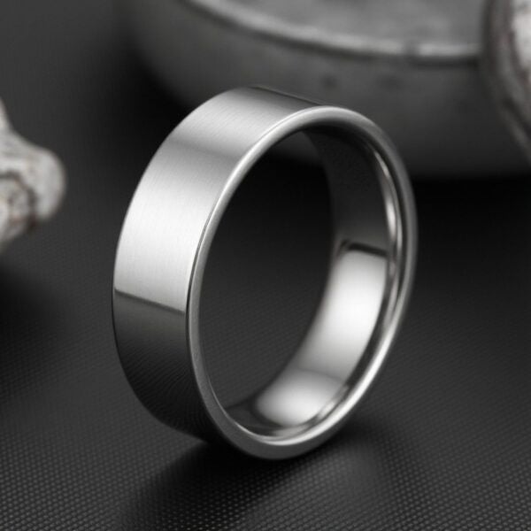 With a grey minimalist background, a ring stands upright. It is a silver tungsten ring that is flat.