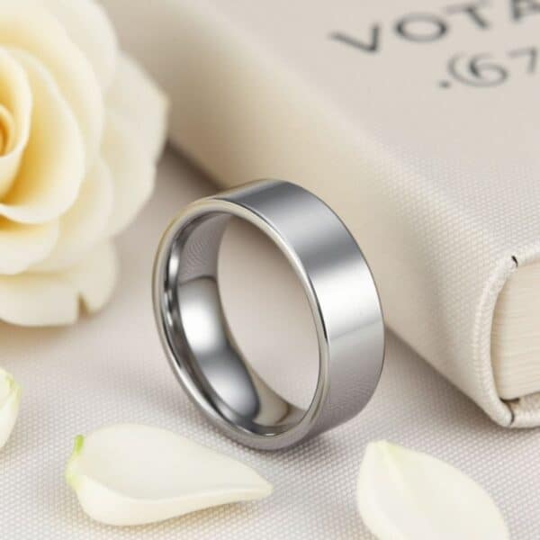 A perfect wedding scene, the image shows a silver wedding band, a white rose and a white wedding vows journal. The ring is a flat silver tungsten ring.