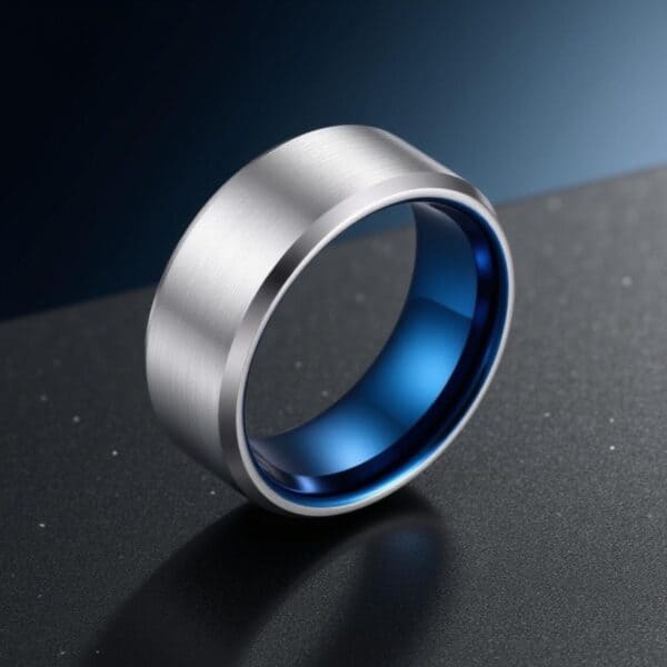 A macro shot of a ring with a dark grey and blue minimalist background. The ring is made with tungsten. It has a flat brushed silver surface and polished silver edges. The inside of the ring is distinctive polished blue.