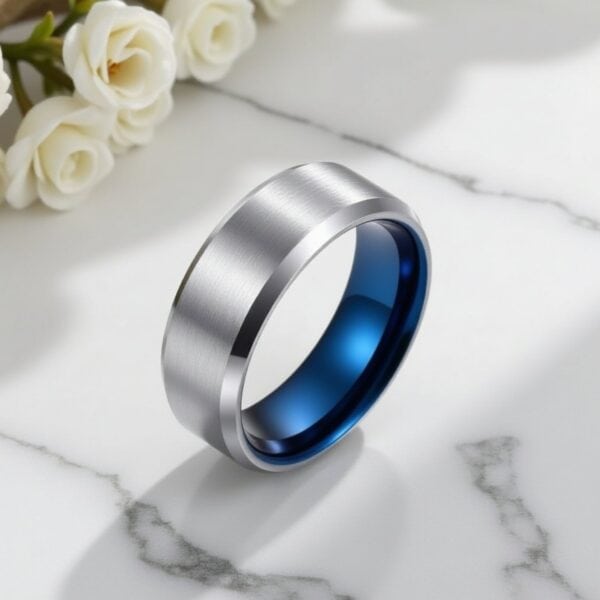 White wedding flowers are in the top corner of the photo. The main feature is a ring which is standing upright on a white marble surface. The ring is a silver tungsten ring with a blue inside. With a mix of brushed and polished finishes.