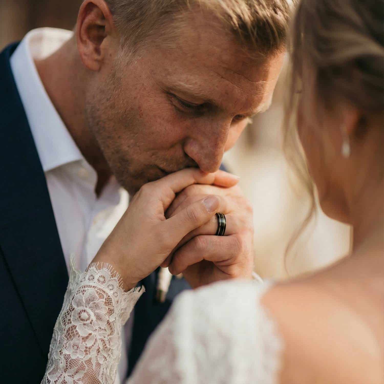 During the wedding ceremony, the groom kisses the brides hand. The man is wearing a black tungsten ring which has distinctive grooves of gold in a double groove.