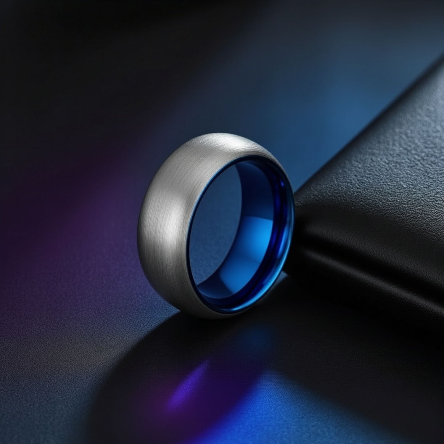 With a ambient iridescent background, a silver tungsten ring stands upright. It has a lightly brushed finish and is slightly domed in shape. The ring has a striking blue interior.
