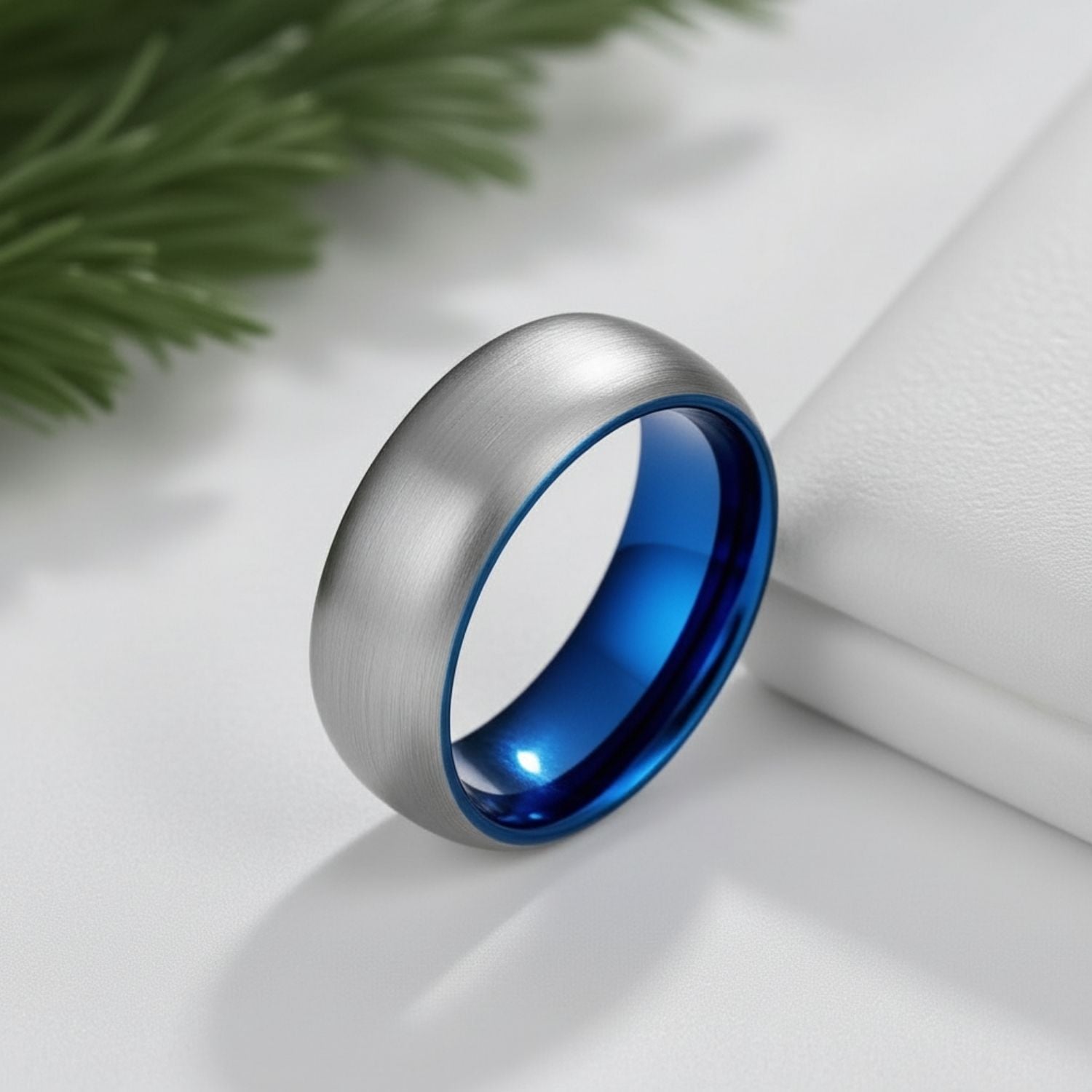 On a white background and with green leaves in the background, a silver tungsten ring stands upright. The ring has a brushed finish and has a polished silver inside.