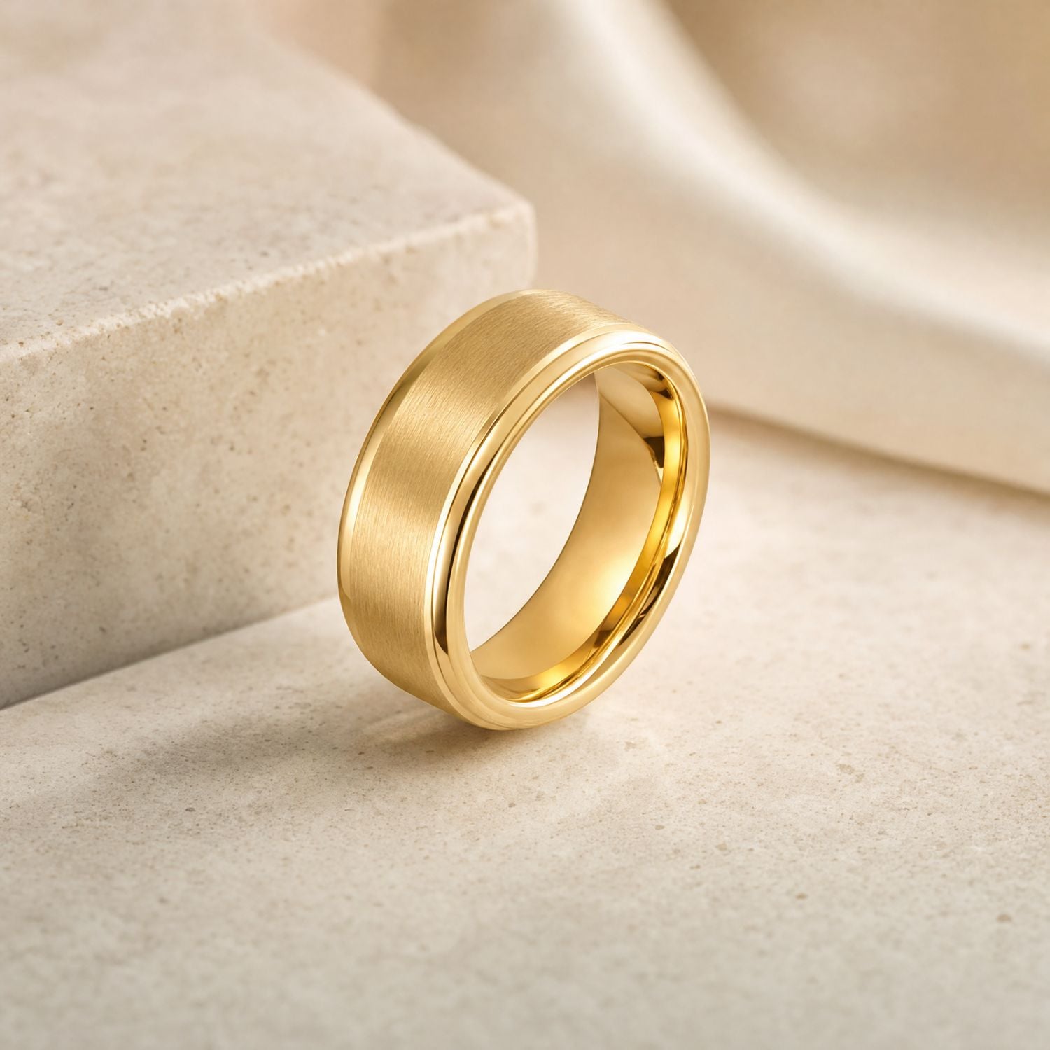 Gold tungsten ring with a brushed centre and polished edges standing upright on a smooth white surface, with a white stone block and soft silk fabric in the background.