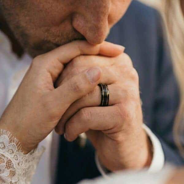 Showing the hands of a newly wed couple, the mans ring is visible, showing a black tungsten ring with a double groove of gold.