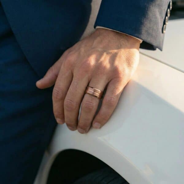 Leaning on the side of a white car, a man wears a navy blue suit. On his hand, he wears a brown and rose gold tungsten ring on his wedding finger.