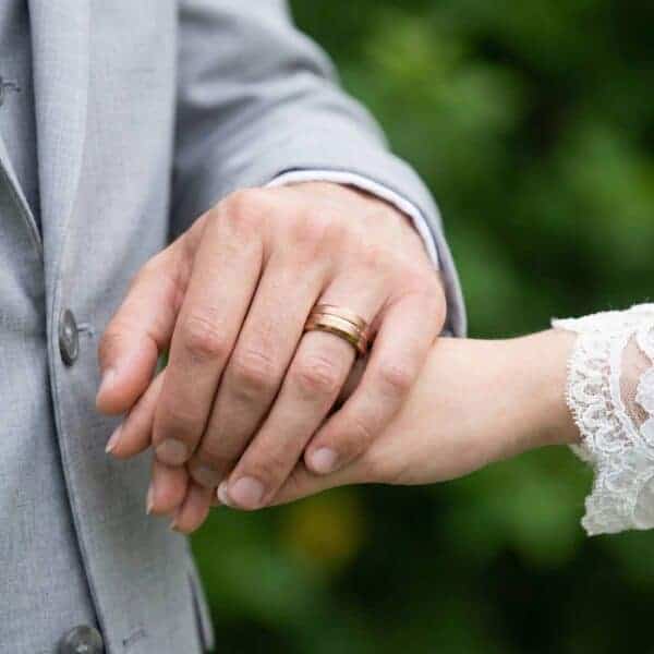 The newlyweds join hands, ready for a photograph. The man's ring is visible. It is a brown tungsten ring featuring a rose gold groove and contrasting brushed and polished finishes.