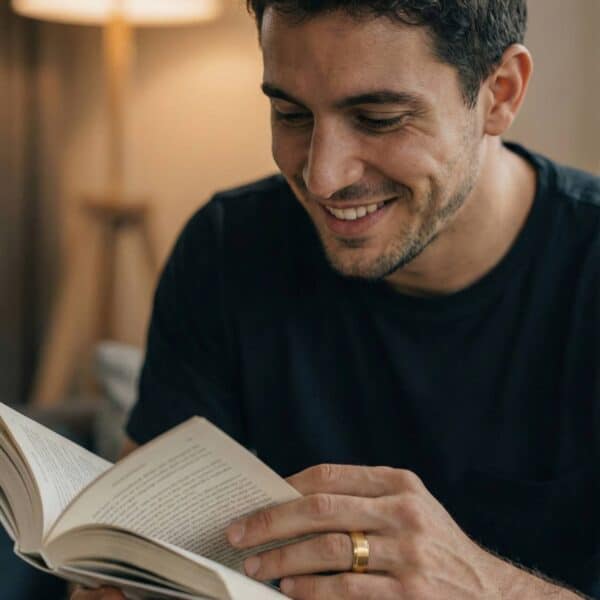 Smiling while he reads, a man enjoys reading his book. Wearing a black top, he wears a gold tungsten ring on his wedding finger.