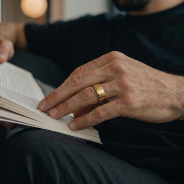 A close up shot of a man reading a book. His hand is ready to turn the page. He is wearing a gold tungsten ring on his finger.
