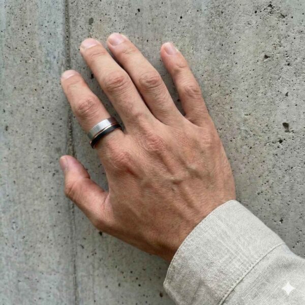 A man rests his hand against the wall. The close up shot shows that he is wearing a tungsten ring that is made up of dual colours - brushed silver, rose gold, and black.
