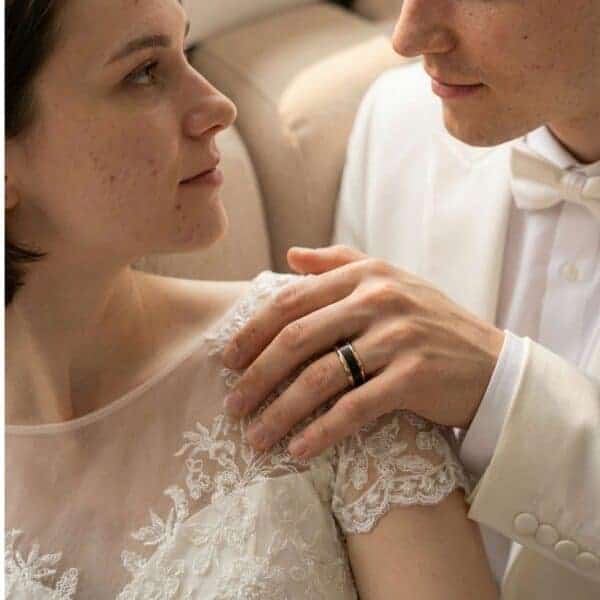 A quiet moment is shared between the newly married couple. The man rests his hand on his partners shoulder. His ring is visible. It is a black tungsten ring with rose gold edges.