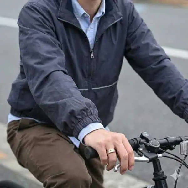 Riding on a bike, a man holds onto the handlebars. Wearing a raincoat, the man has a gold and silver tungsten ring on his hand.
