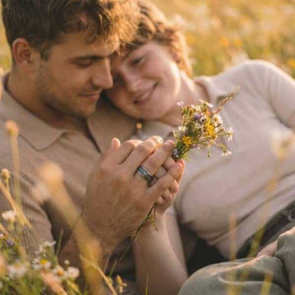 A blissful scene of a happy couple relaxing on the long grass in the countryside. Smiling and leaning against each other, the man holds a small bunch of yellow flowers. On his finger, he wears a silver tungsten ring that features inlays of natural wood, and abalone shell.
