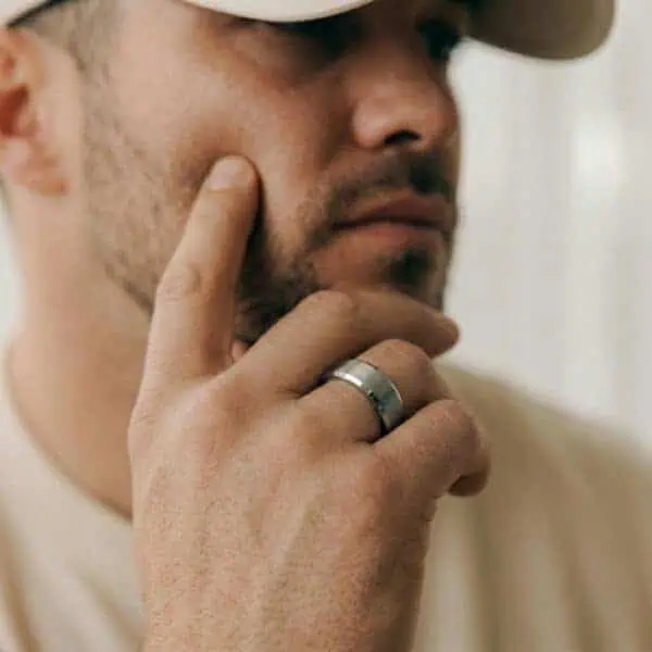 A close up of a mans face who is wearing a matching cream t-shirt and cap. He has his hand held up to his face. He is wearing a silver tungsten ring. It has polished bevelled edges and a brushed centre section.