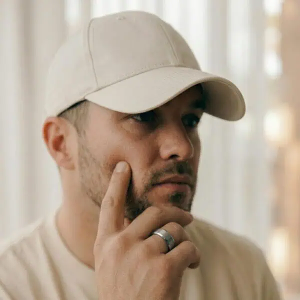 A man looks lost in thought, wearing a cream cap and a matching cream t-shirt. He has his hand held up to his chin. He is wearing a silver tungsten ring that has a brushed centre and polished bevelled edges.