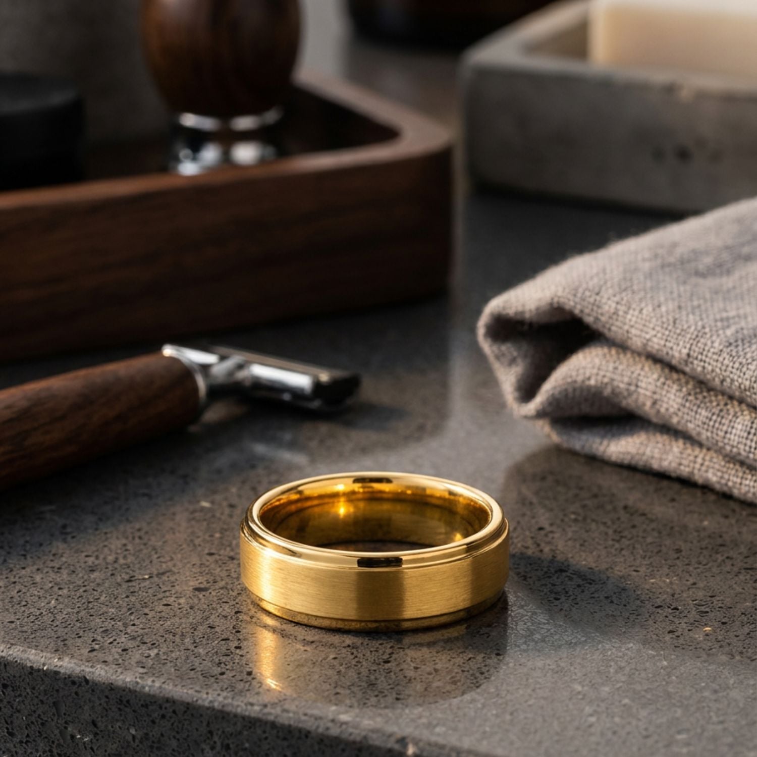Men's gold tungsten ring with a brushed centre lying on its side on a grey marble-effect bathroom countertop beside a grey flannel and shaving accessories.