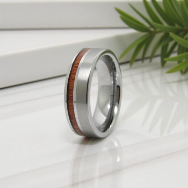 On a smooth white marble worktop, green leaves in the background, a silver tungsten ring sits in the foreground. The ring features a natural wood inlay.