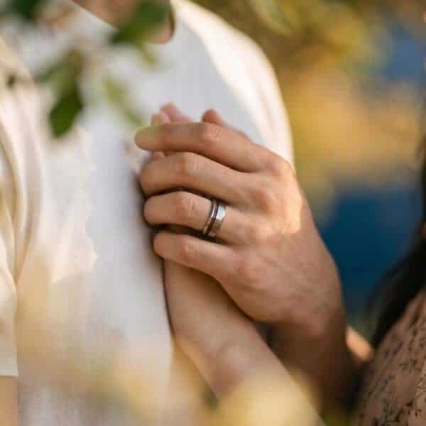 A newly married couple join hands. The bride wears a white wedding gown and the hands are held up near her dress. The man is wearing a silver tungsten ring that has a groove of wood running through it.