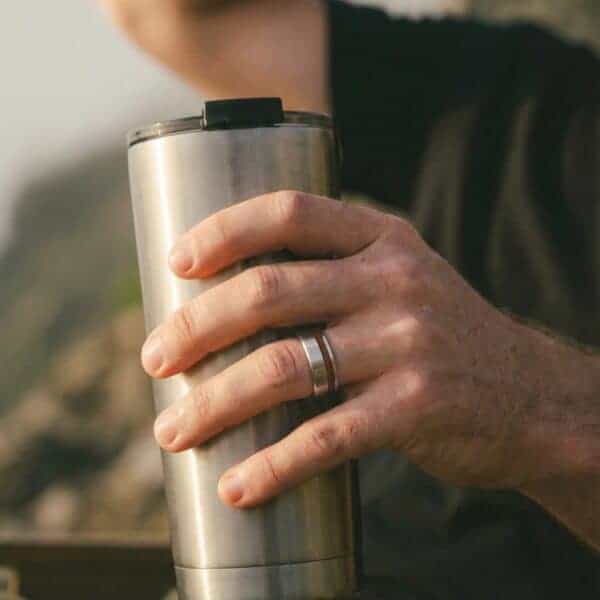 Holding a coffee in a travel mug, you can see a close up of a mans hand. He is wearing a silver tungsten ring that has a groove of natural wood.