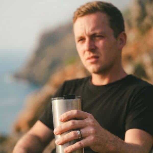 Enjoying the glorious coastal view and the sunshine, and man sits in a thoughtful pose. He is holding a silver travel mug and is wearing a silver tungsten ring which has a groove of wood.