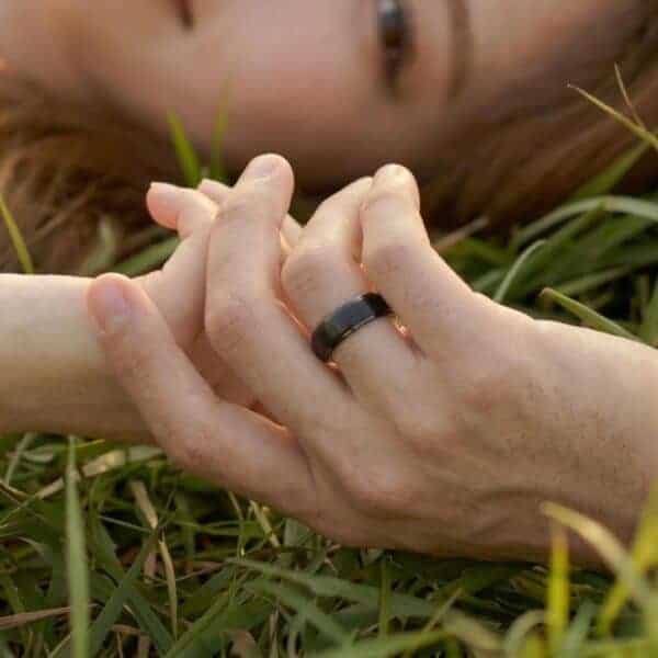A couple lay on the grass holding hands. You can see a small part of the face of the woman. The couple are holding hands. The man is wearing a black tungsten ring. The centre of the ring is raised and has a brushed finish.