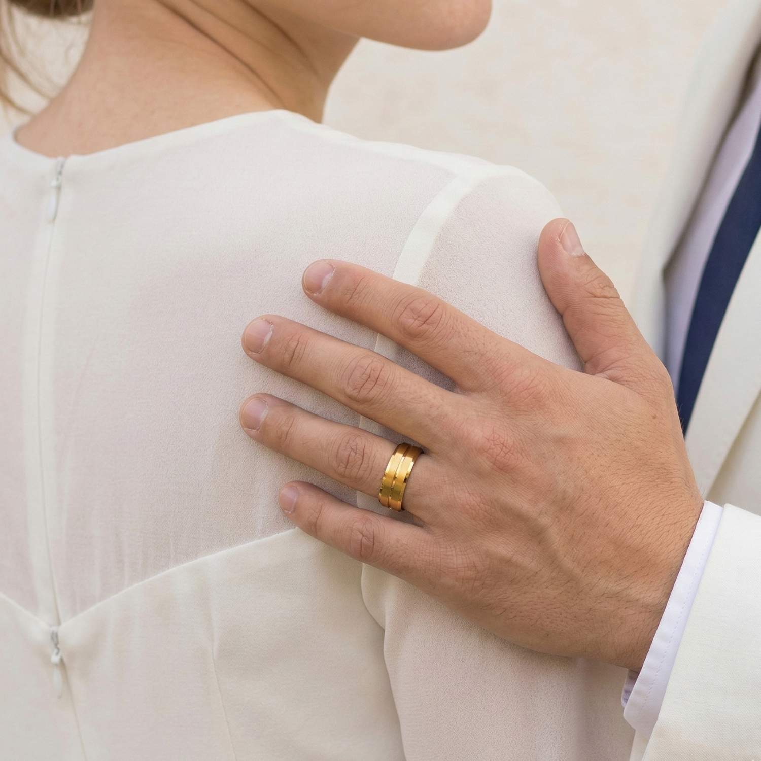A newly married couple stand close together. The man rests his hand on his partners shoulder. He is wearing his new wedding band which is a gold titanium ring featuring a centre groove and bevelled edges.