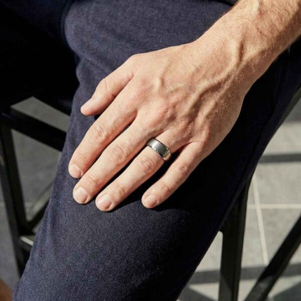 With his hand resting on his leg, the mans ring is on display. It is a silver tungsten ring. The brushed centre section of the ring contrasts with the polished bevelled edges. The inside of the ring is a vivid red.