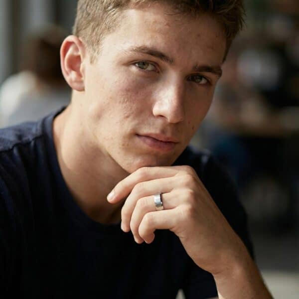 Looking into the camera, a young man sits looking thoughtful with his hand resting under his chin. On his finger, he wears a silver tungsten ring. The interior of the ring is vivid red.