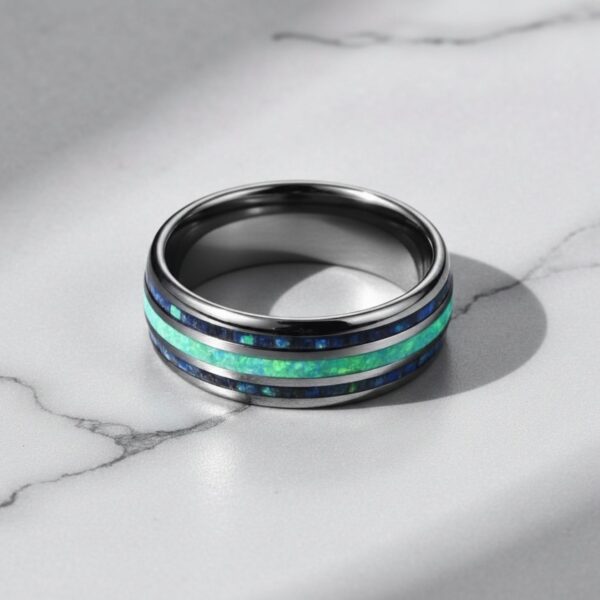 On a white marble shelf, a ring lays flat. It is a silver tungsten ring which has grooves of abalone shell and turquoise.