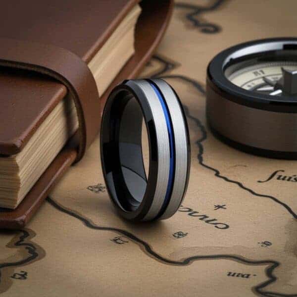Set amongst a leather bound book,. a compass, and a map, a tungsten ring stands upright, ready for travel and adventure. The ring has two raised sections which are brushed silver. A groove of vivid blue runs through the centre of the ring. The bevelled edges of the ring are polished black, and the inside of the ring is black.