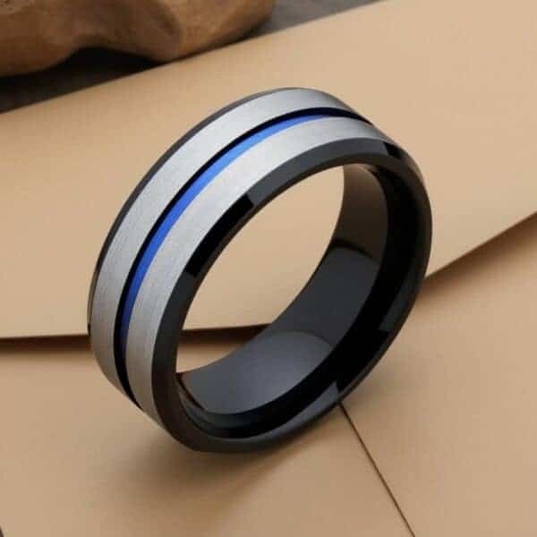 A close up image of a silver tungsten ring featuring a blue groove and polished black edges. The ring is upright, on top of a brown envelope.
